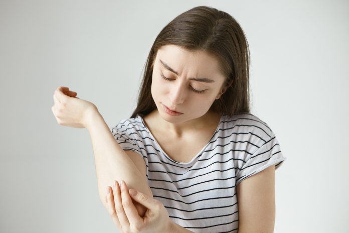 Studio picture of frustrated young dark haired female studying skin on her arm after she fell off bike. Student girl dressed in stylish striped t-shirt looking at her elbow, feeling itch or pain